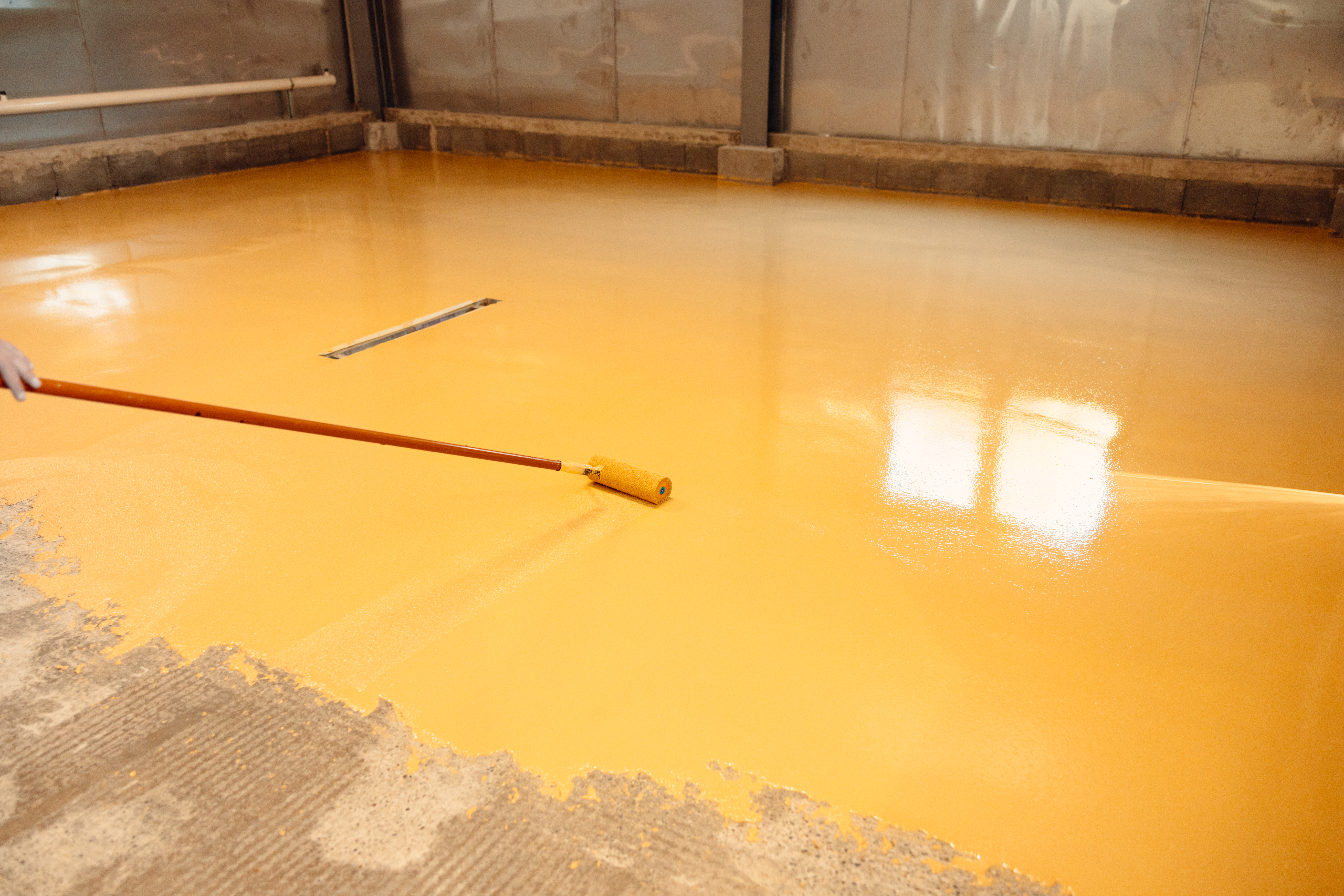 Construction worker is applying epoxy resin on a large concrete floor with a roller to create a resistant and waterproof surface